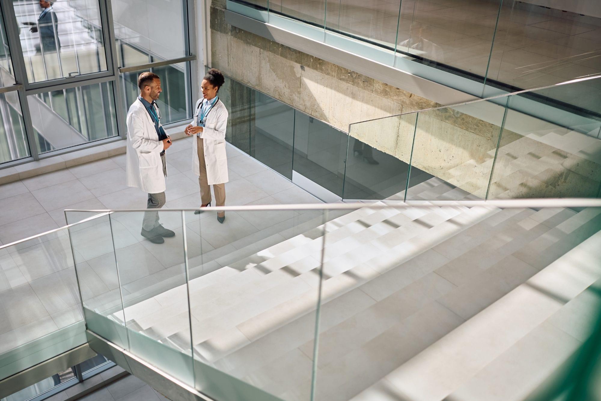 Two doctors talking on a hospital atrium landing beside a glass staircase