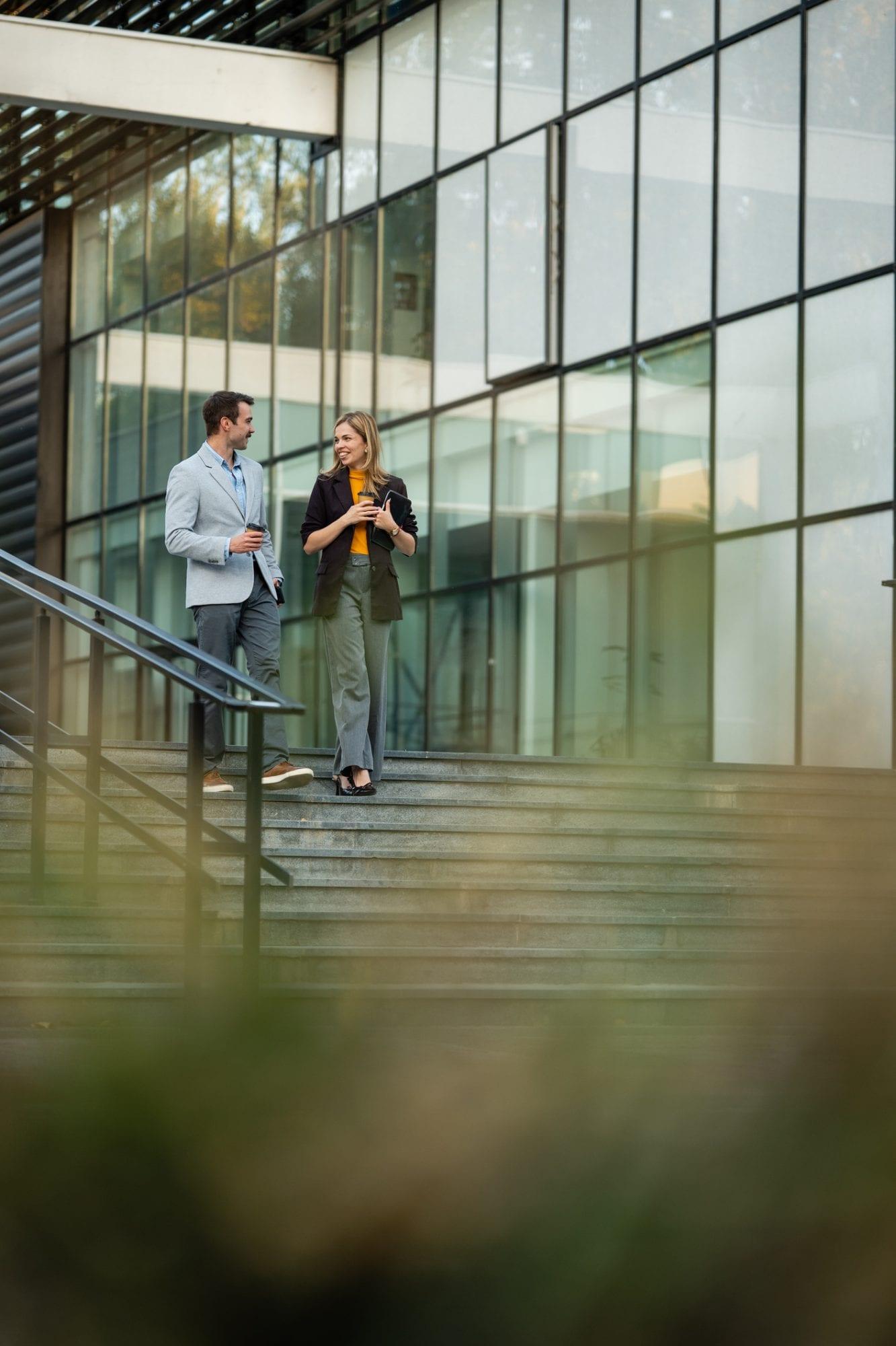 Two colleagues with coffee talking as they walk down the steps of a glass office building