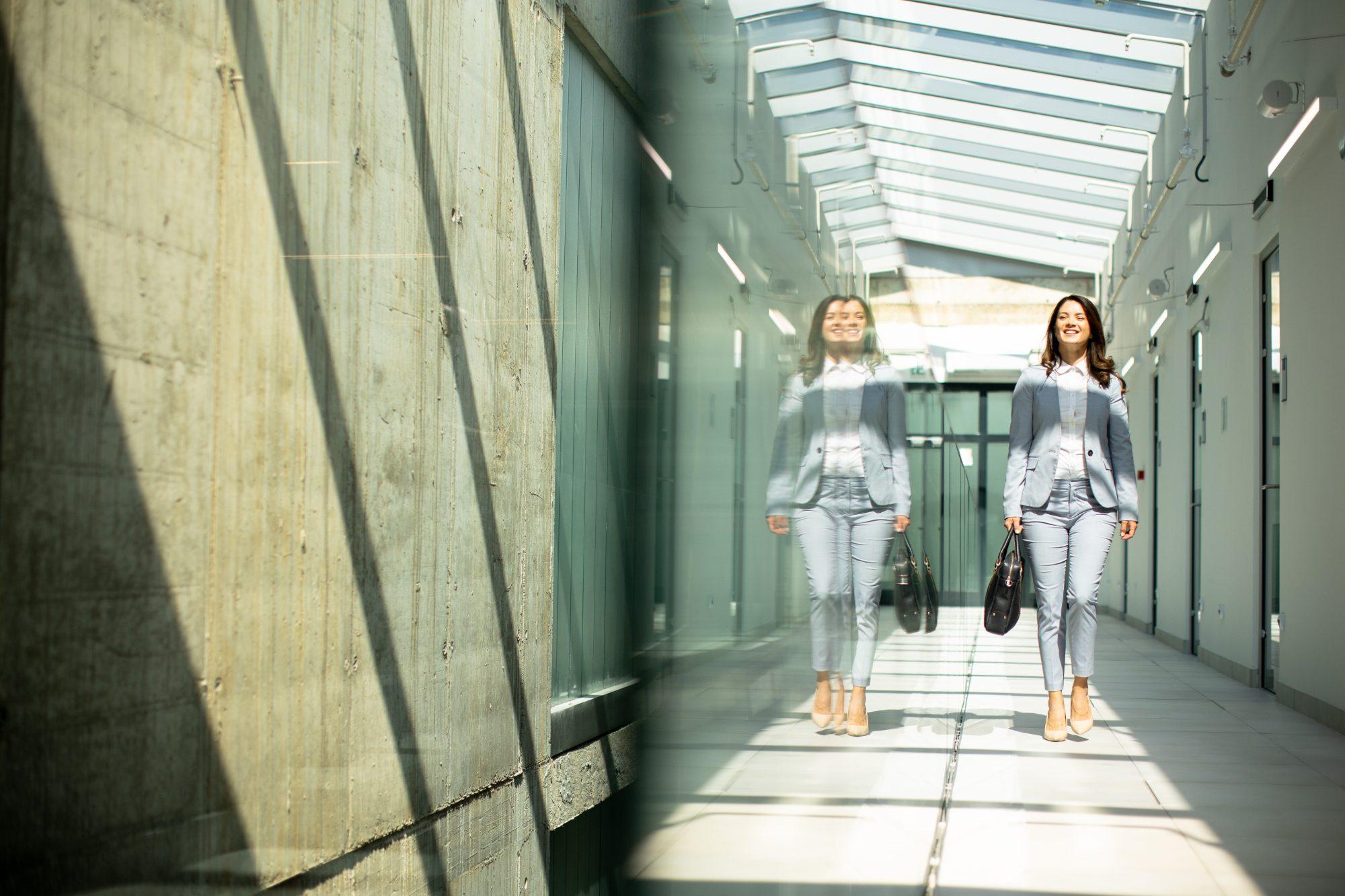 Smiling woman in a light grey suit carrying a briefcase along a sunlit glass-roofed corridor, with her reflection in the wall beside her