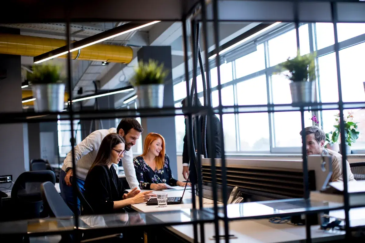 A team gathered around a laptop in an open-plan office, seen through a metal shelf