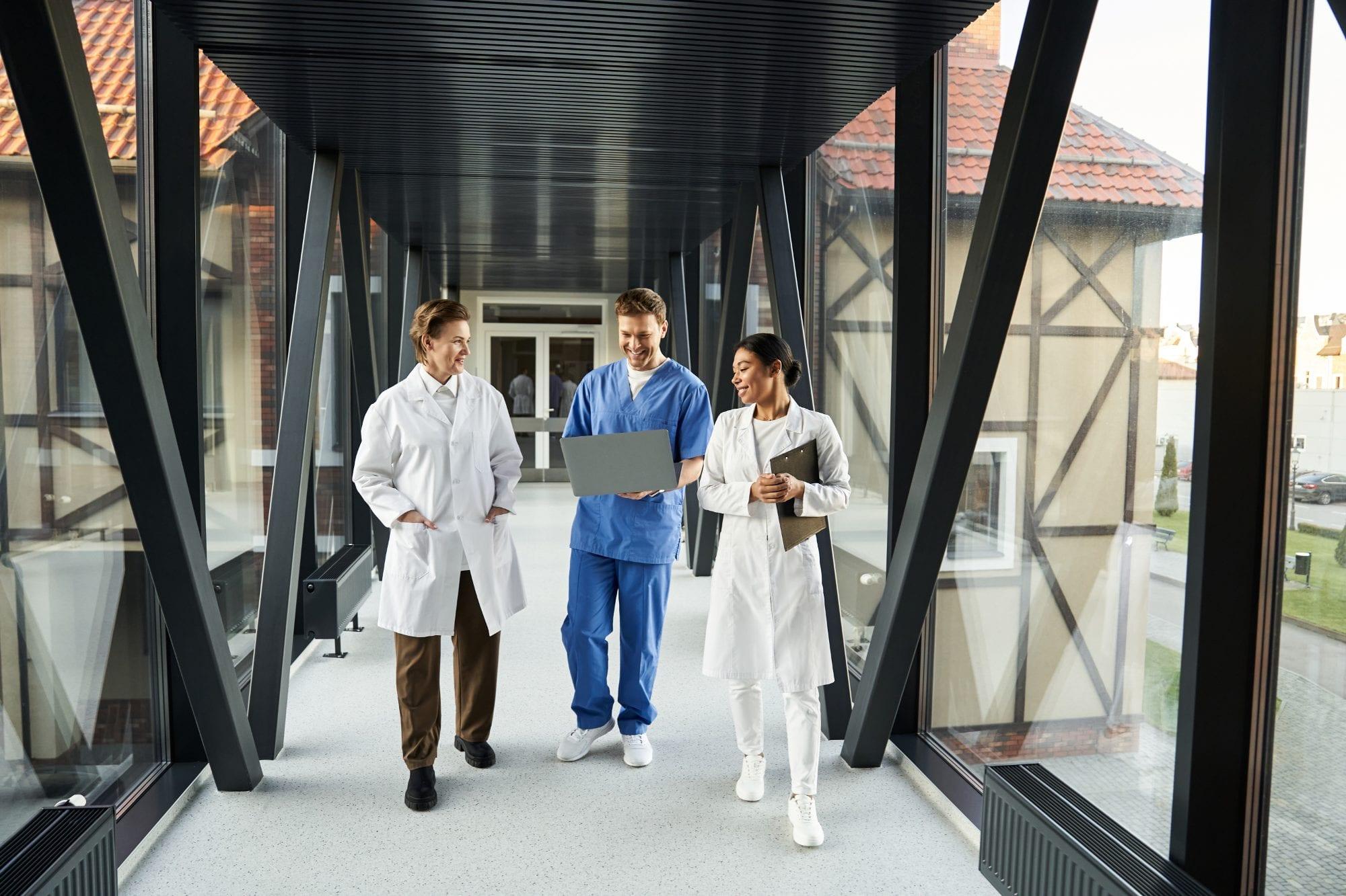 Three clinicians reviewing a laptop as they walk through a glass hospital walkway