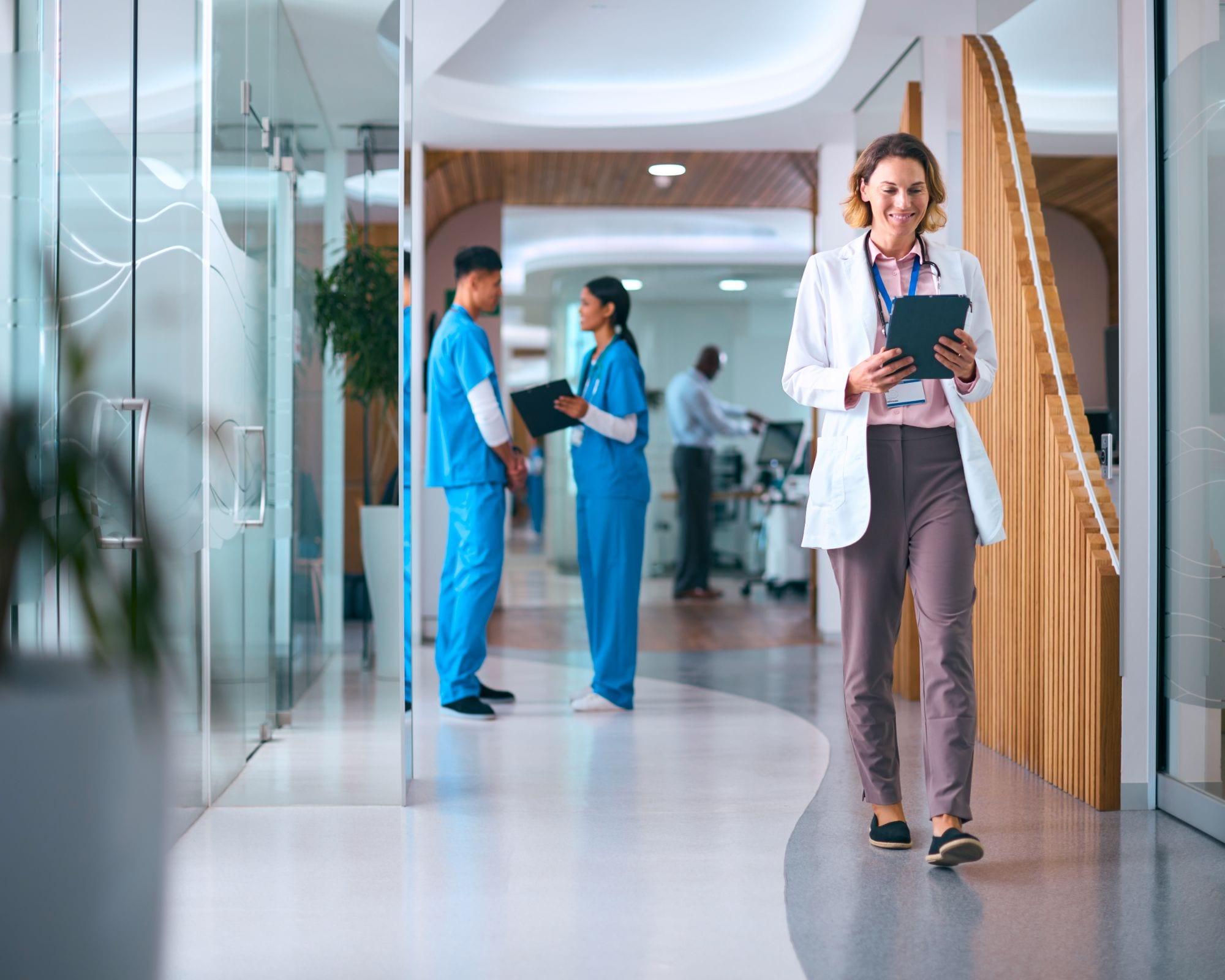 Doctor in a white coat reviewing a tablet while walking through a hospital corridor
